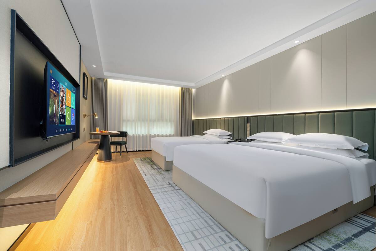 A modern hotel room with a large flat-screen TV mounted on the wall, a wooden floor, two beds with white bedding, a small table, and a window with curtains.