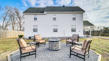 A white house with a covered porch, a gravel patio with four chairs and a round table, and a wooden fence in the background.