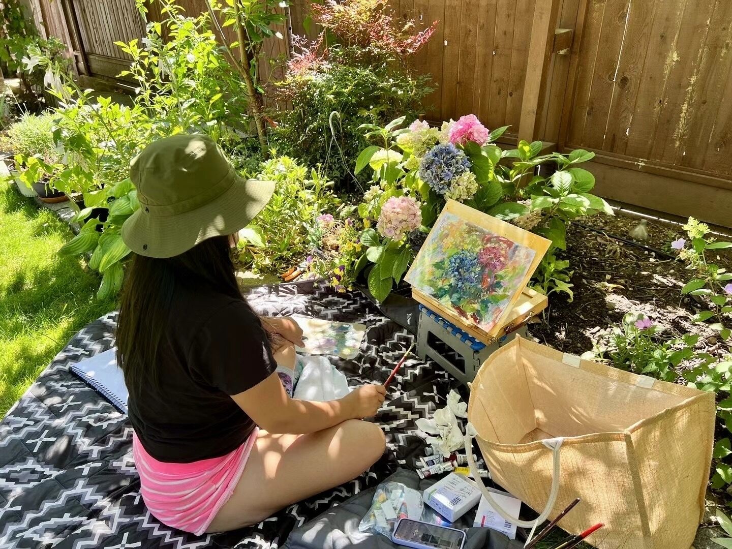 Une personne peignant en plein air, assise sur un tapis devant un chevalet pliable, entourée de différentes plantes et fleurs.