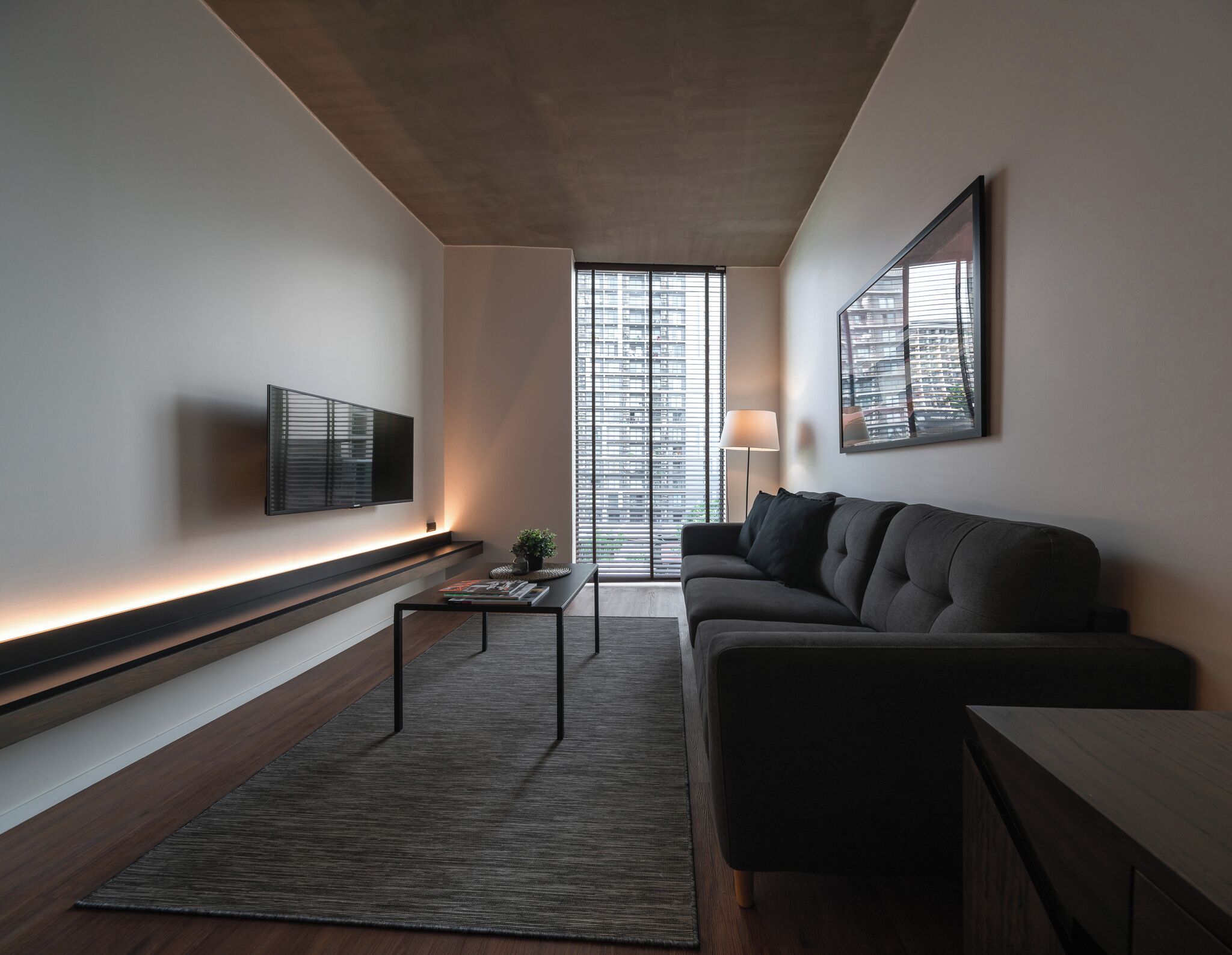 Un salon moderne avec un canapé, une table basse, un téléviseur fixé au mur et de grandes fenêtres équipées de stores.