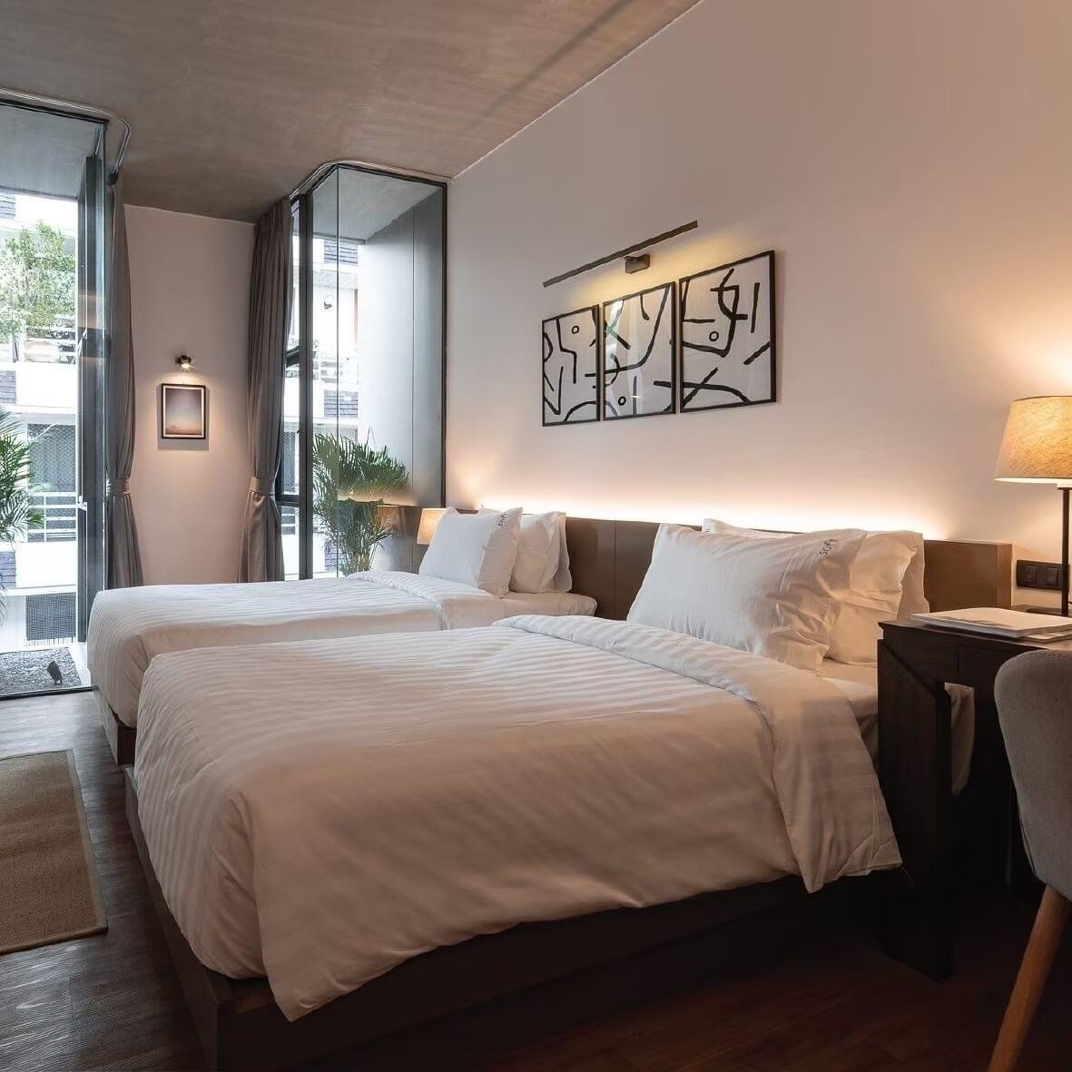 A hotel room with two beds, a large window, a wall-mounted artwork, and a bedside table with a lamp.