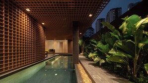 Un área interior de piscina moderna con techo de diseño enrejado, una piscina larga y un camino bordeado de guijarros y plantas tropicales.