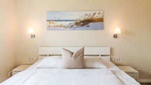 Una cama bien hecha con sábanas blancas y una sola almohada color beige, con dos mesitas de noche y lámparas a ambos lados. Encima de la cama hay un cuadro de una playa.