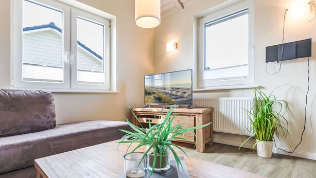 Una sala de estar moderna con un sofá, una mesa de centro con objetos decorativos, un televisor y ventanas grandes con persianas.