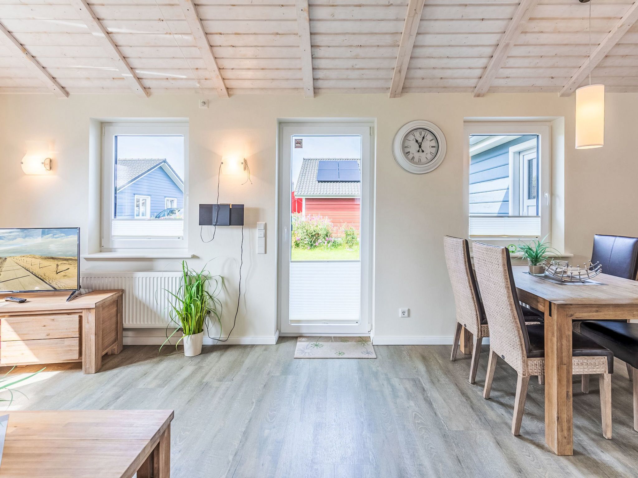 Ein moderner, gut beleuchteter Raum mit Holzbalkendecke, einer Essecke mit Tisch und Stühlen, einem Fernseher auf einem Holzbodenständchen und Blick auf ein Haus durch ein Fenster.