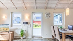Una habitación moderna y bien iluminada con techo de madera, una zona de comedor con mesa y sillas, un televisor sobre un mueble de madera, y vistas a una casa a través de una ventana.