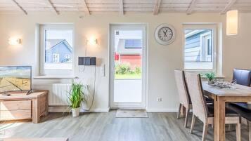 Ein moderner, gut beleuchteter Raum mit Holzbalkendecke, einer Essecke mit Tisch und Stühlen, einem Fernseher auf einem Holzbodenständchen und Blick auf ein Haus durch ein Fenster.