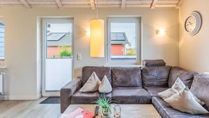 Een moderne woonkamer met een bruine leren bank, een houten salontafel en een rode map. Er zijn twee ramen met uitzicht op een rood gebouw en een groen landschap.
