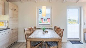 Een eetgedeelte met een houten tafel en stoelen, een raam met uitzicht op een huis, en een keuken met houten kastjes.
