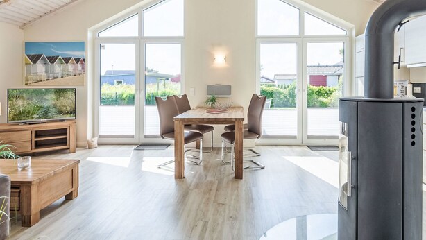 A spacious room with a wooden dining table, two chairs, a TV on a wooden stand, and a large window offering a view of greenery and buildings. - Luv & Lee by Interhome (Dagebüll)
