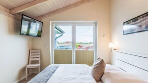 Hotellihuone, jossa on sänky, seinälle asennettu televisio, tuoli ja ikkuna, jossa on sälekaihtimet.
