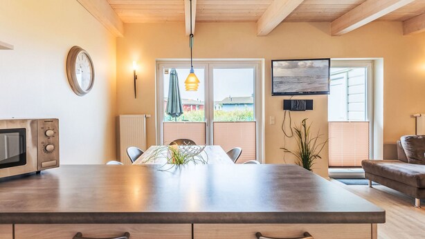 A modern kitchen with a large island, wooden ceiling beams, a dining area with chairs, a television, and a view of the outdoors. - Nordseeluft by Interhome (Dagebüll)