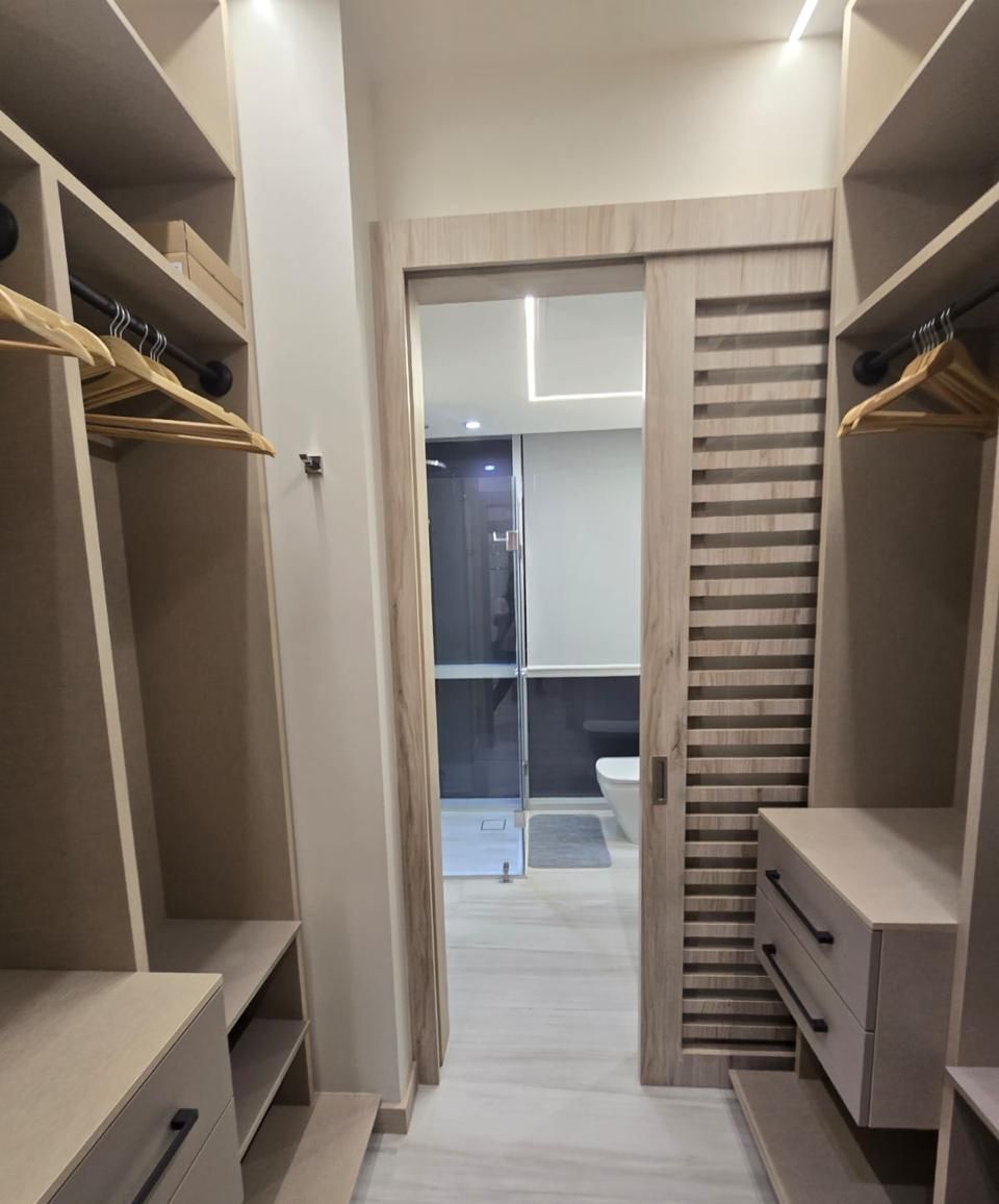 Un vestidor con estanterías abiertas, una puerta corredera que da al baño y un panel divisor de listones de madera.
