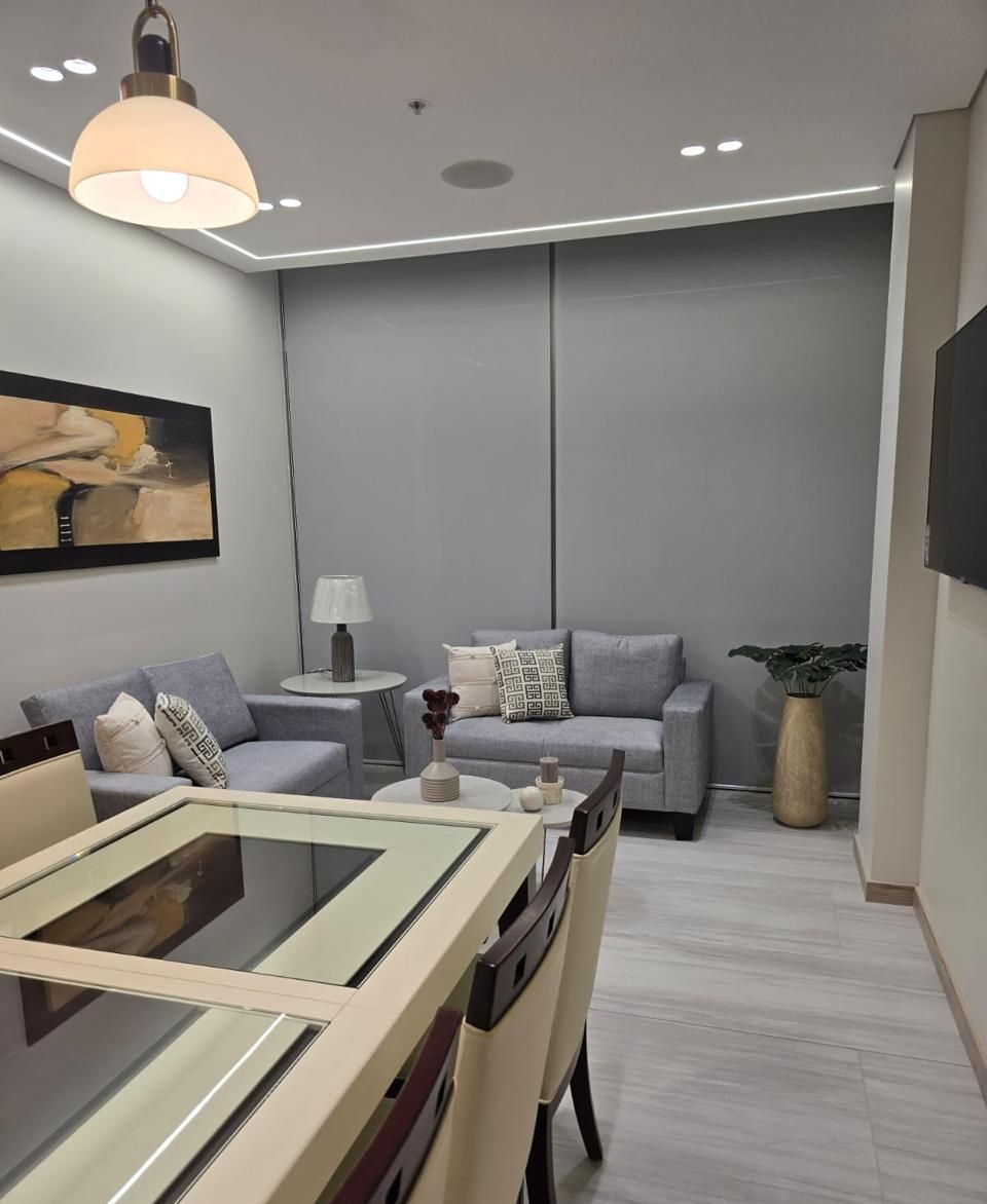 Una sala de estar moderna con una mesa de comedor de vidrio, sofás grises, una mesita de noche con lámpara y una planta grande en maceta.