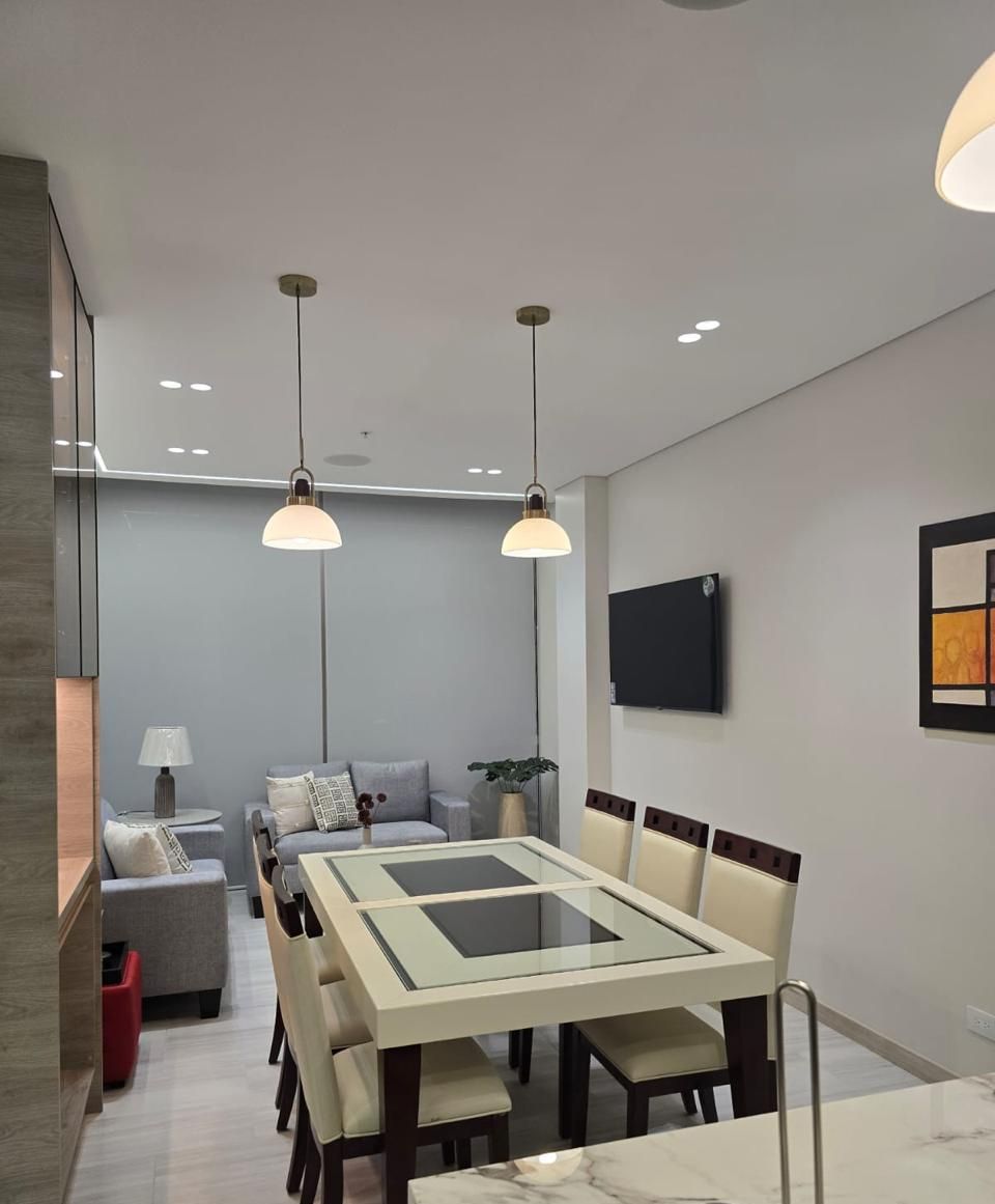 Un comedor moderno con una mesa de vidrio, sillas, luces colgantes y un televisor de pantalla plana en la pared.