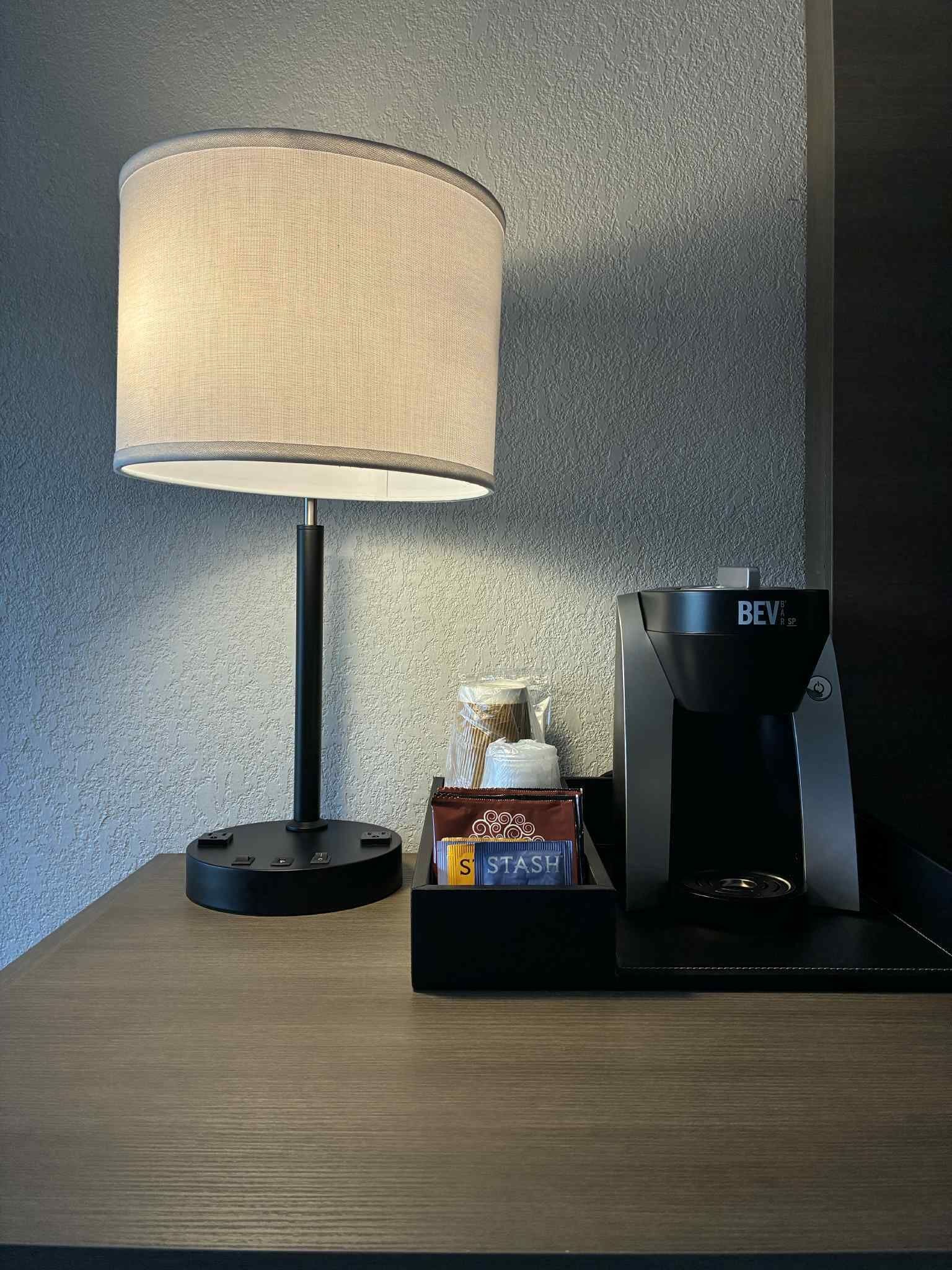 Una lámpara, una cafetera y una caja con la etiqueta 'STASH' sobre una superficie de madera frente a una pared con textura.