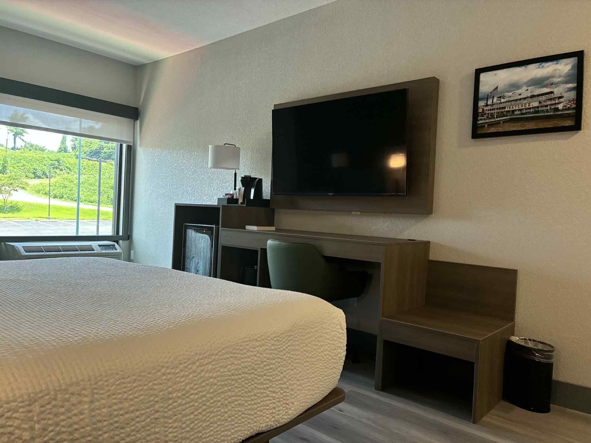 Une chambre d’hôtel avec un grand lit, un bureau avec un ordinateur, un téléviseur fixé au mur et une fenêtre donnant sur un paysage verdoyant.