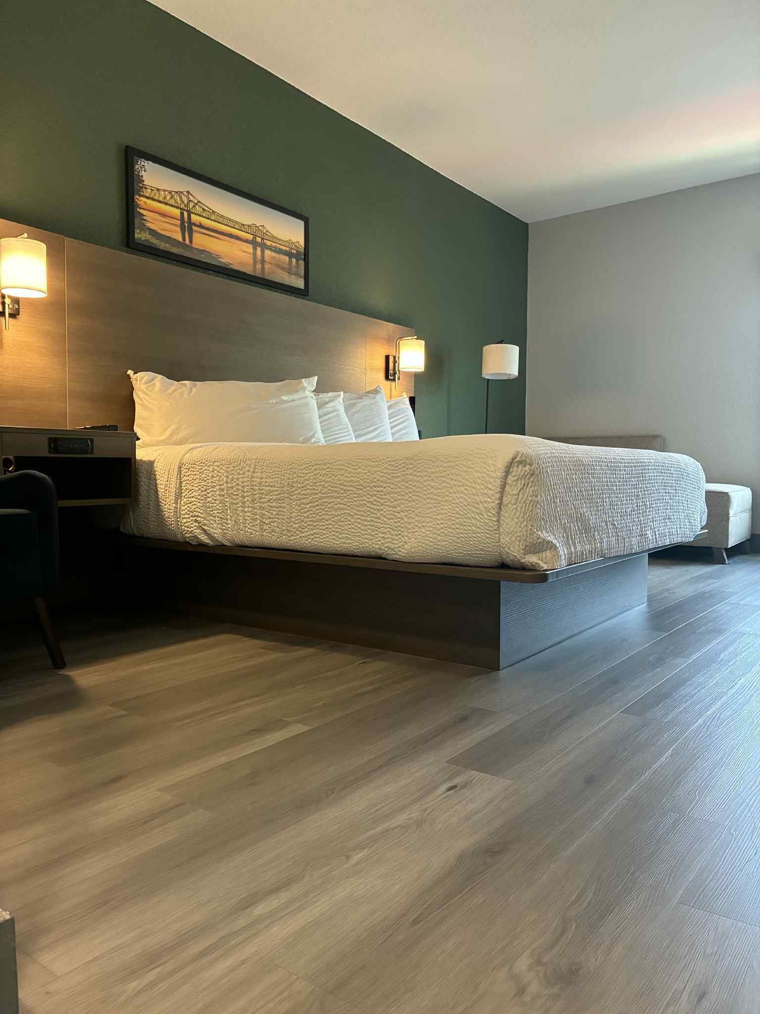 A modern hotel room with a large bed, a headboard with a painting, bedside lamps, and a wooden floor.