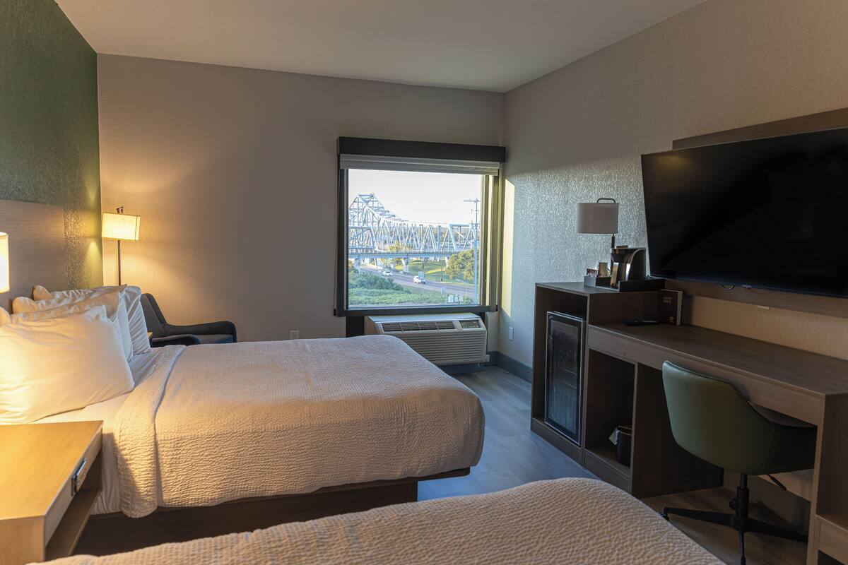 A hotel room with two beds, a desk, a TV, and a window with a view of a bridge.