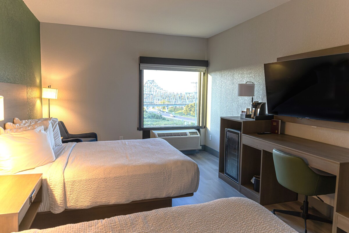 A hotel room with two beds, a desk, a TV, and a window with a view of a bridge.