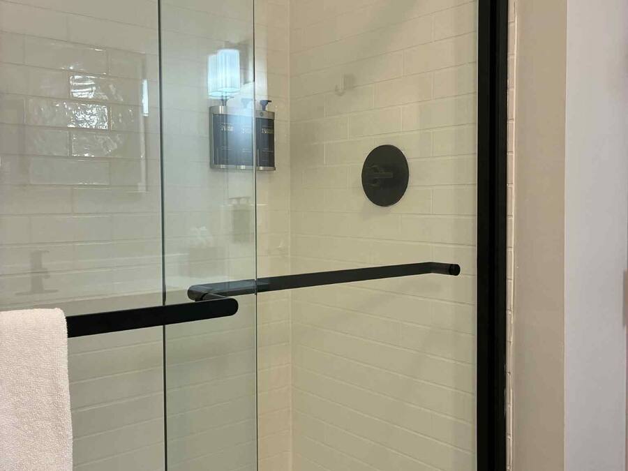 Cabina doccia in vetro con cornice nera, pareti rivestite di piastrelle bianche e un asciugamano appeso alla porta.