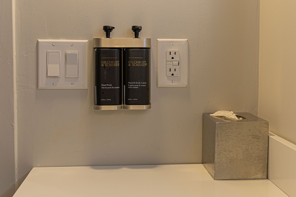 A bathroom with a hand and body lotion dispenser, a tissue box, and a light switch.