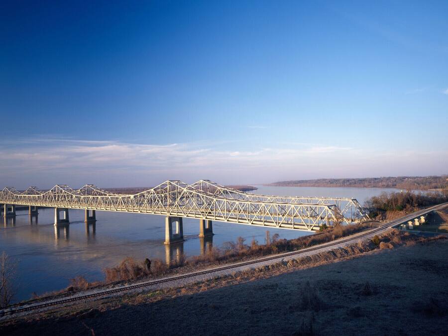 Un pont d’acier enjambe une rivière paisible, avec une voie ferrée qui longe la rivière.