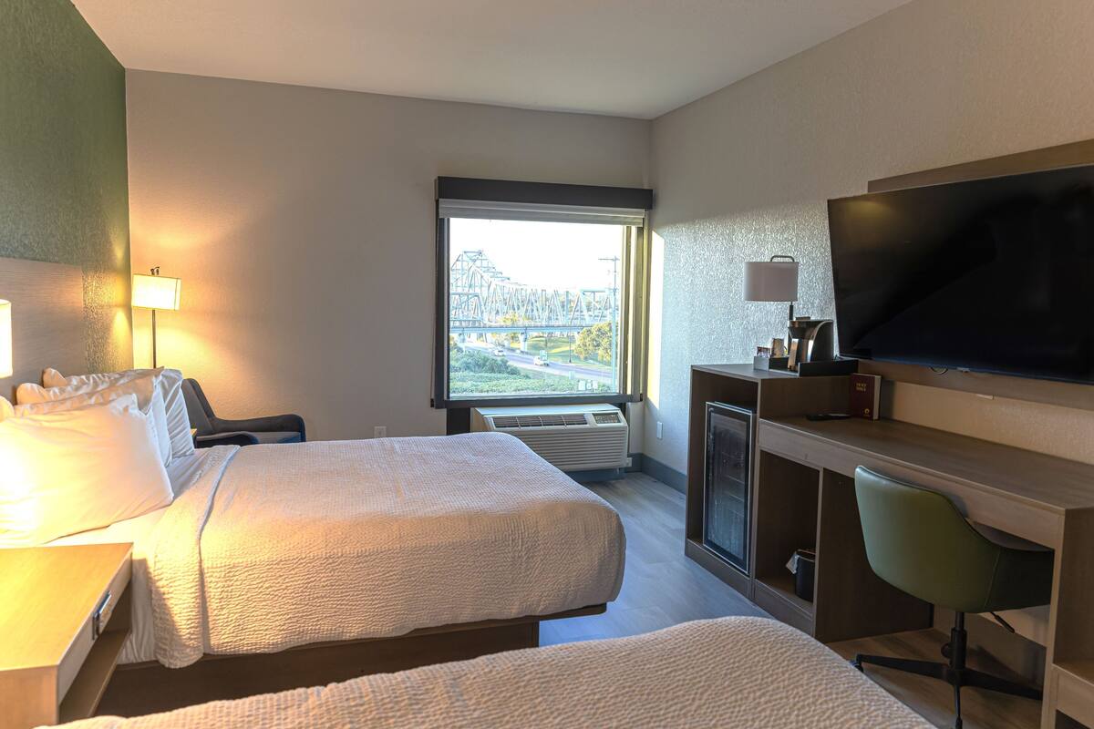 A hotel room with two beds, a desk, a TV, and a window with a view of a bridge.