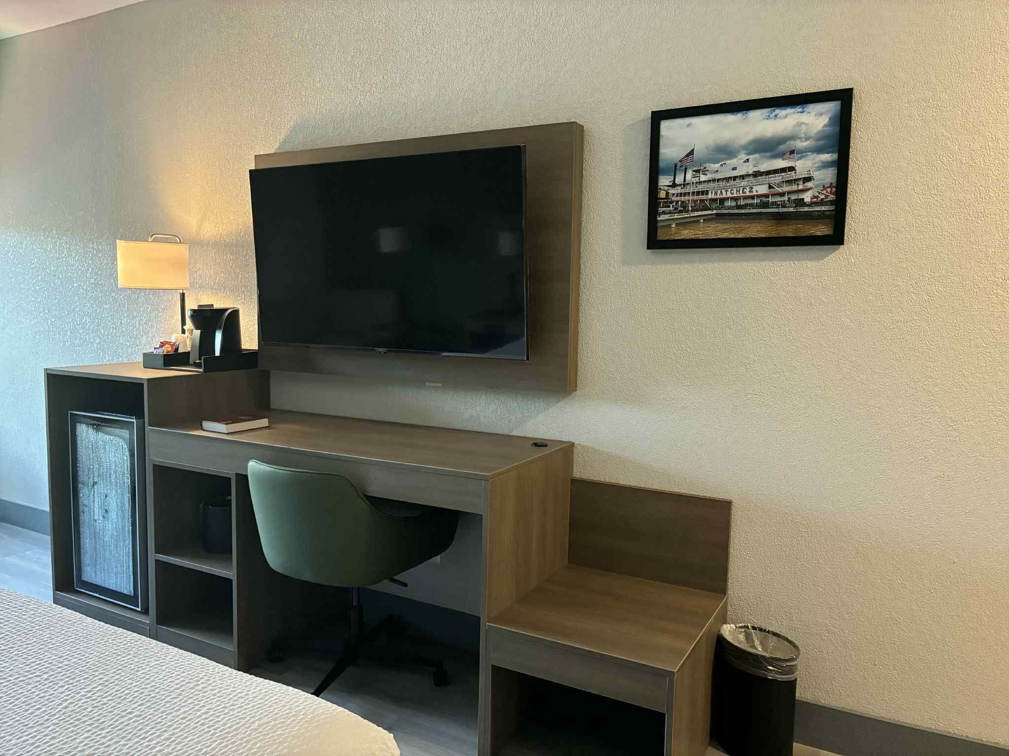 Habitación de hotel con televisión de pantalla plana montada en la pared, escritorio con silla, lámpara y un cuadro con una imagen de un barco.