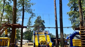 An outdoor playground with inflatable structures, including a slide and climbing net, surrounded by trees.