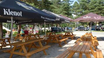 Mesas de picnic y sombrillas al aire libre con la marca 'Klenot', ubicadas en un parque con árboles y coches estacionados al fondo.