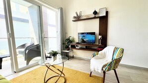 Una sala de estar moderna con una mesa de vidrio, una silla colorida, un televisor de pantalla plana y un balcón con puerta corrediza.