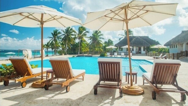 Un área junto a la piscina con sillones de descanso y sombrillas, un cielo azul despejado y palmeras.