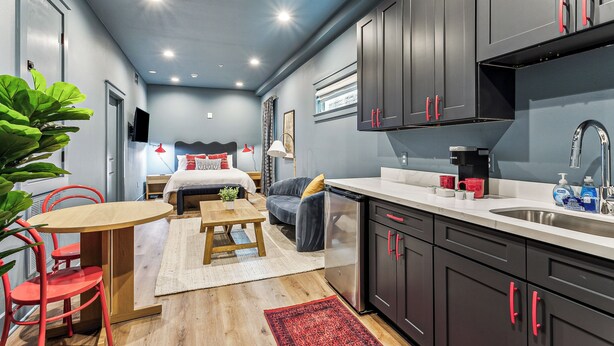 A compact hotel room with a kitchenette, a bed with a headboard, a dining area with a table and chairs, and a large potted plant.