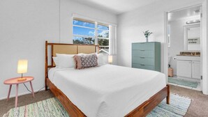 A bedroom with a wooden bed, a window, a bedside table with a lamp, a cabinet, and a bathroom door.