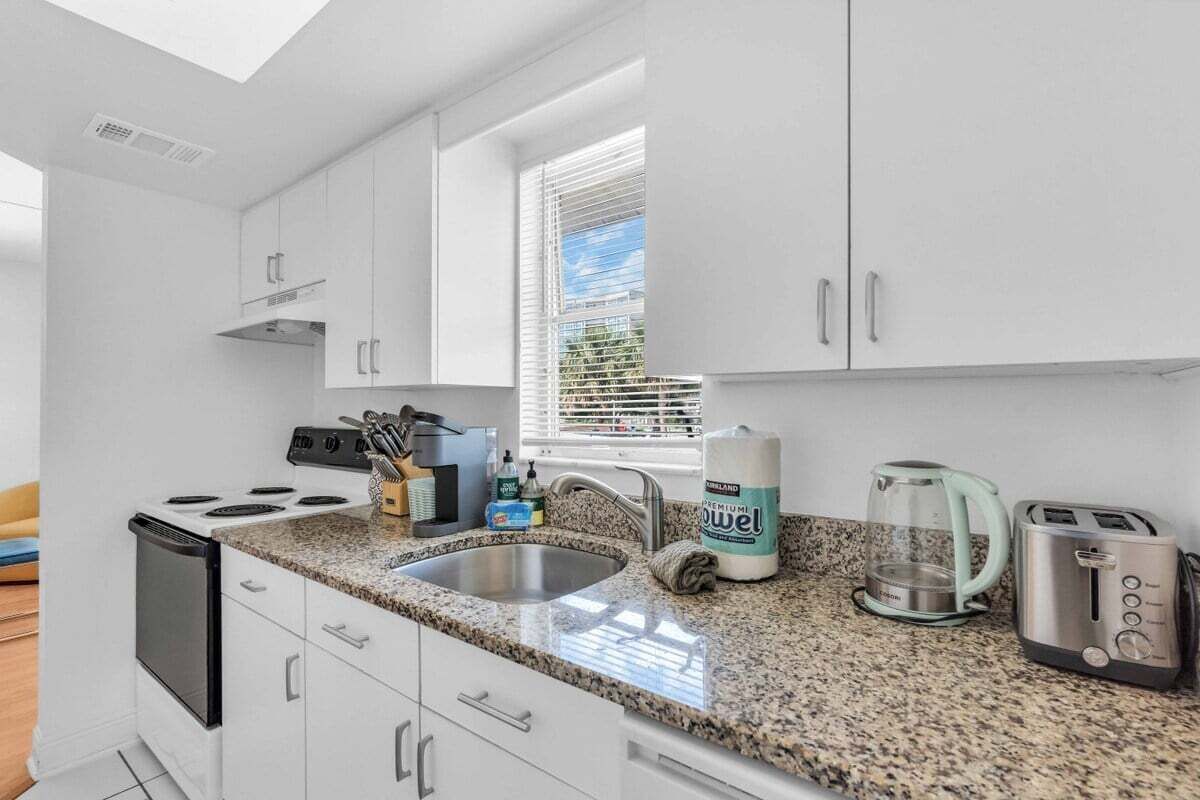 Una cocina moderna con armarios blancos, encimera de granito, estufa, fregadero, cafetera y tostadora.