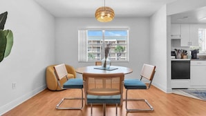 A dining area with a round table, two chairs, a hanging light fixture, and a window with a view of buildings.