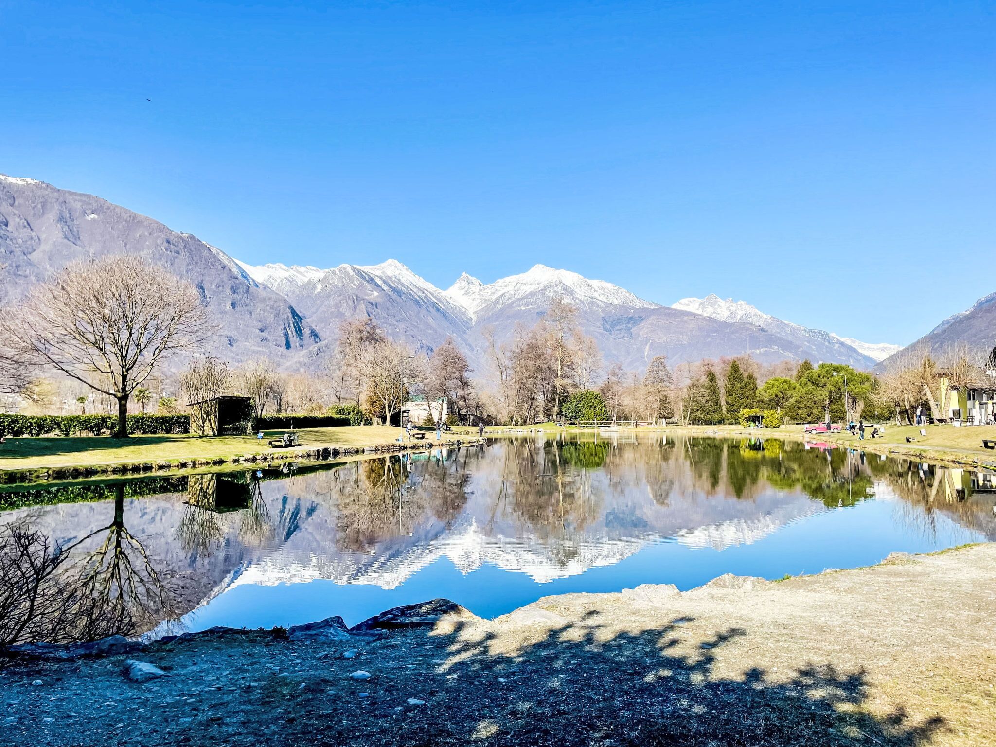 Un lac paisible, avec des montagnes en arrière-plan, entouré d’un parc agrémenté de bancs et d’arbres.