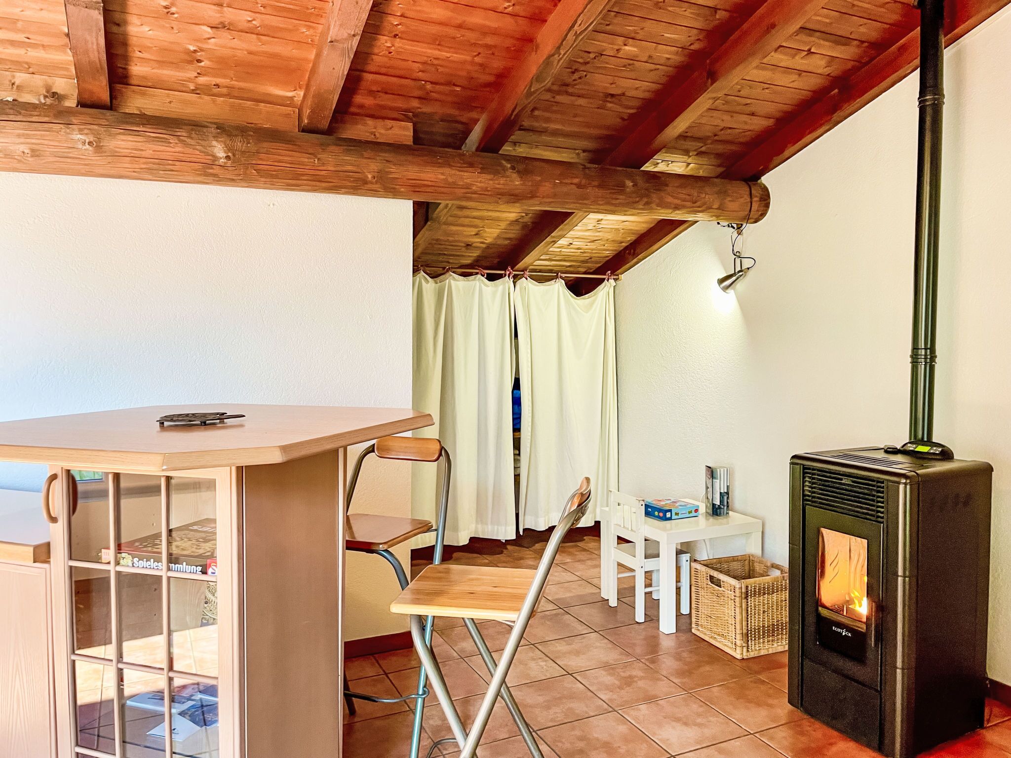 Une pièce chaleureuse avec un plafond en bois, une petite table entourée de chaises, un poêle et une fenêtre avec des rideaux.