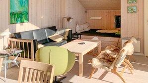 Una habitación acogedora con un sofá, una butaca verde, una mesa de madera y un sillón de piel. Hay un cuadro en la pared y una mesita con una lámpara.