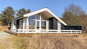 Una casa moderna de un piso con ventanas amplias, una terraza de madera y un techo inclinado.
