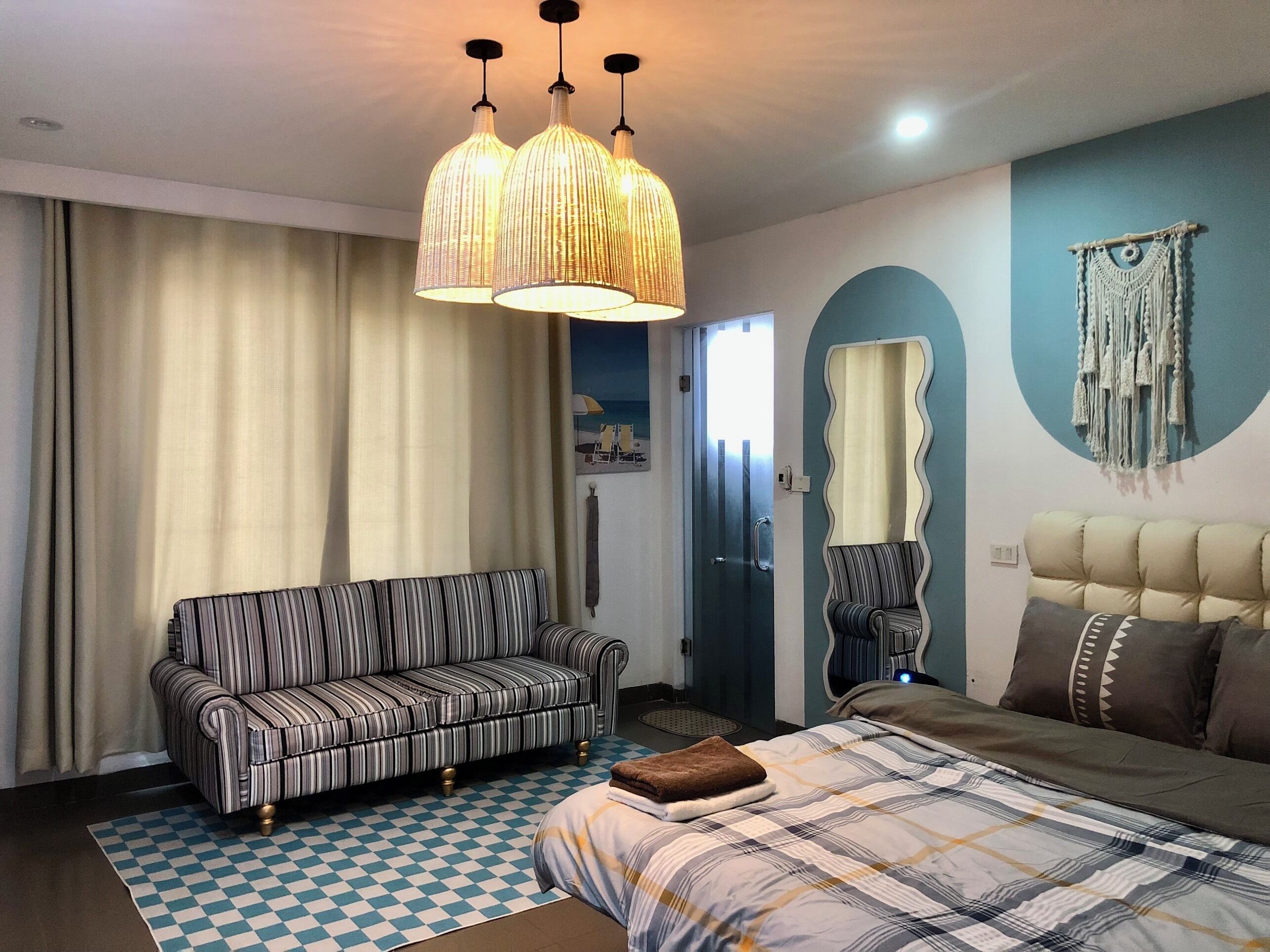 A hotel room with a striped sofa, a bed with a plaid bedspread, a checkered rug, a large mirror, and a macrame wall hanging.