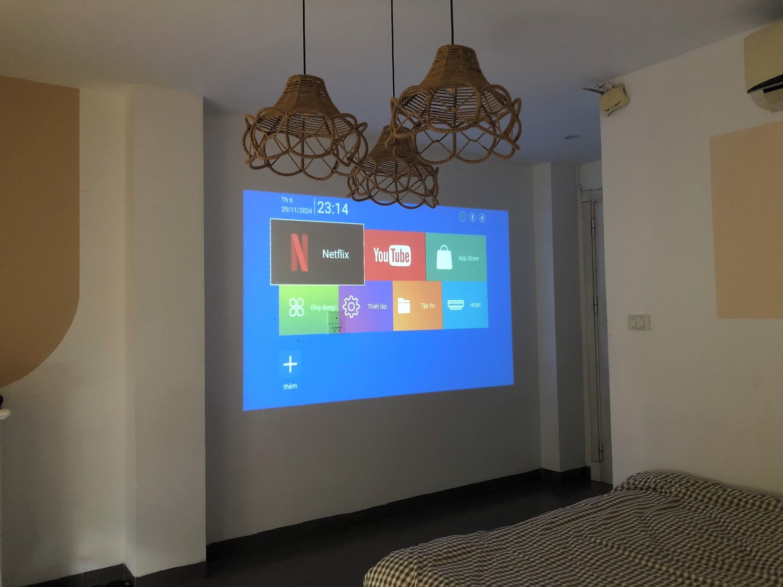 A projection screen displaying a digital interface with app icons, including Netflix and YouTube, in a room with a bed and hanging wicker lamps.
