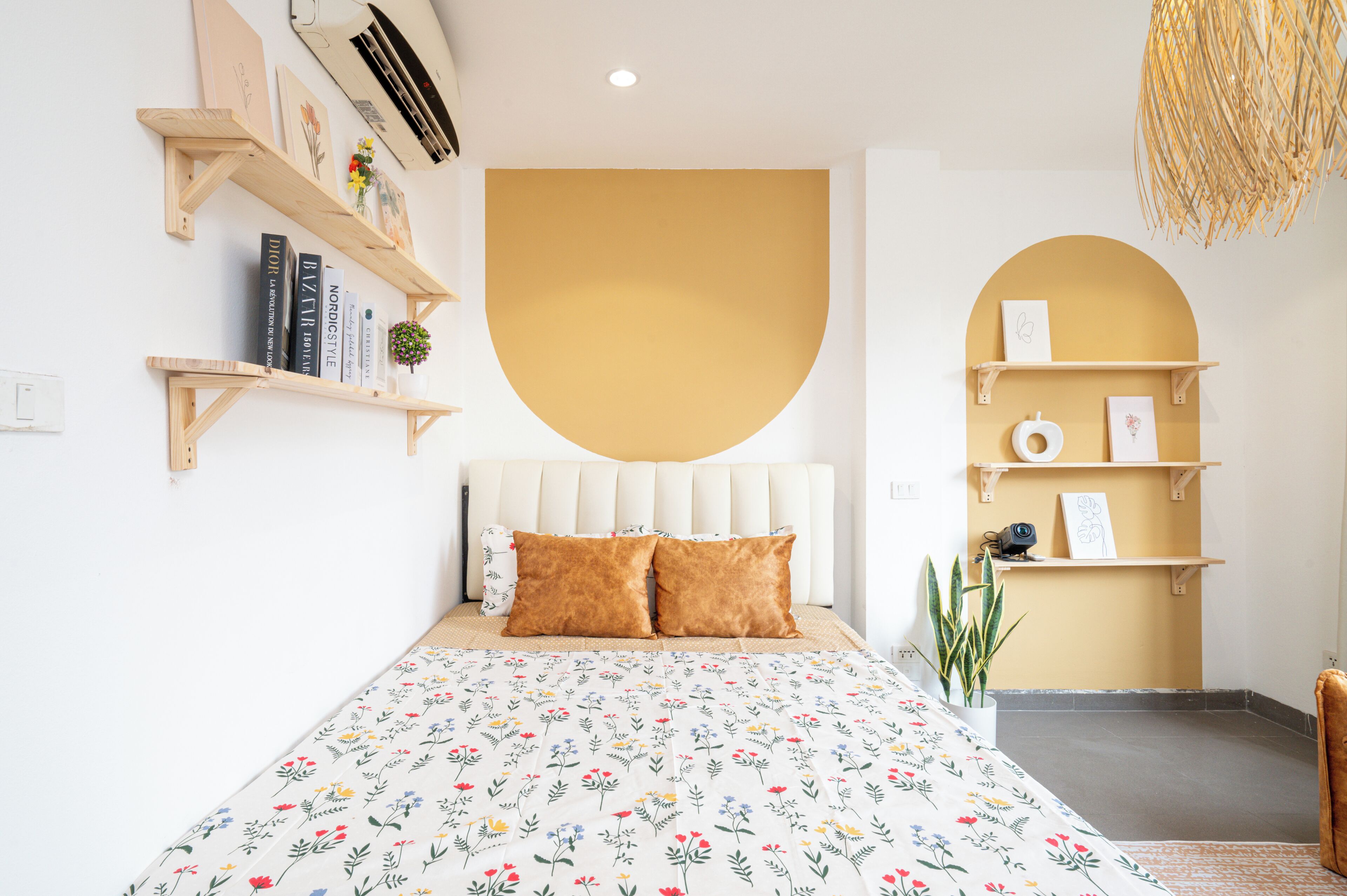 Een slaapkamer met een bed, een wit hoofdeinde, een geel accentmuur en houten planken met boeken en decoratieve objecten.