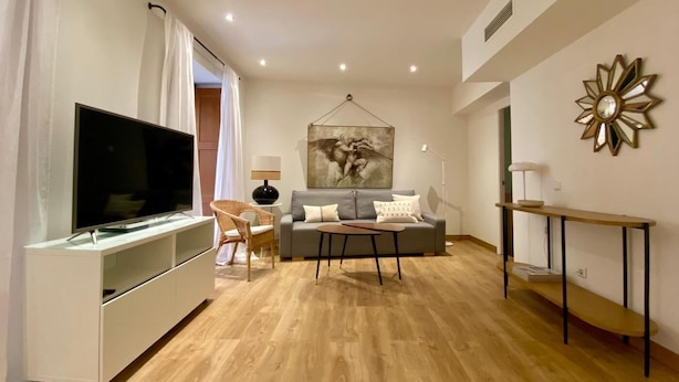 A modern living room with a flat-screen TV, a grey sofa, a wicker chair, and a coffee table.