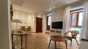 A modern living room with a wooden floor, a dining area with wicker chairs, a flat-screen TV on a white stand, and a large window with curtains.