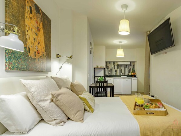 A compact hotel room with a bed, a dining area, a kitchenette, and a TV.