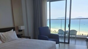 Hotelkamer met een groot bed, een stoel, een nachtkastje en uitzicht op het strand door een glazen deur.
