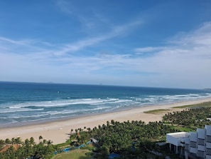 A beach with a view of the ocean, palm trees, and a building with a modern design. - Spring home Arena Cam Ranh (Bac Cam Ranh)