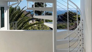 Uma escadaria espiral branca com corrimão de metal, que leva a uma varanda externa com vista desobstruída para palmeiras e uma paisagem distante.